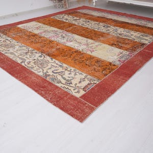 Puede incluir: Una alfombra rectangular con un dise&ntilde;o de patchwork de franjas verticales alternas. El patr&oacute;n incluye secciones de tela color crema con intrincados motivos florales en tonos apagados y paneles de color naranja amarronado, todo enmarcado por un borde rojo ancho.