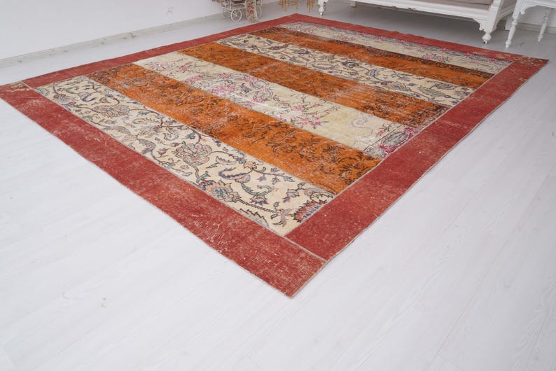 Puede incluir: Una alfombra rectangular con un dise&ntilde;o de patchwork de franjas verticales alternas. El patr&oacute;n incluye secciones de tela color crema con intrincados motivos florales en tonos apagados y paneles de color naranja amarronado, todo enmarcado por un borde rojo ancho.