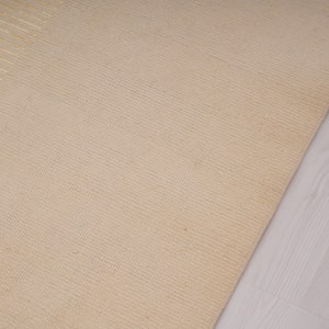 Pu&ograve; includere: Primo piano di un tappeto beige tessuto con un sottile motivo testurizzato. Il tappeto &egrave; realizzato in fibre naturali e ha una sensazione morbida e naturale.