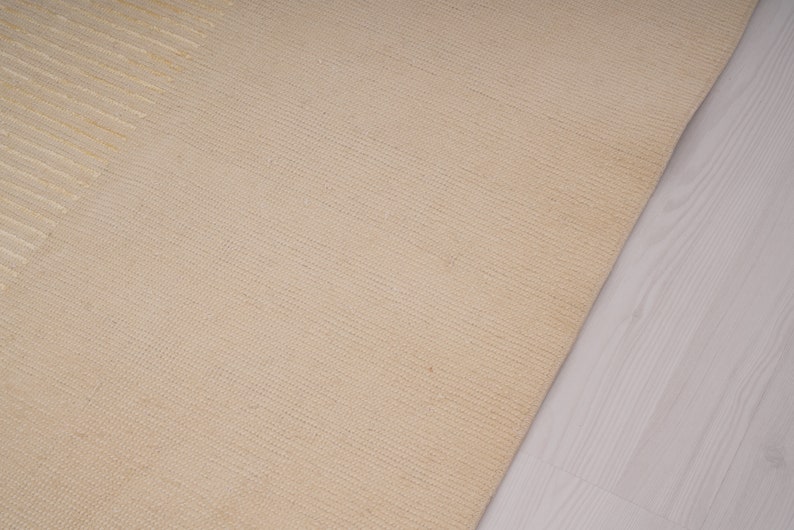 Pu&ograve; includere: Primo piano di un tappeto beige tessuto con un sottile motivo testurizzato. Il tappeto &egrave; realizzato in fibre naturali e ha una sensazione morbida e naturale.