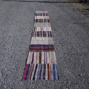 May include: A long, narrow runner rug featuring a patchwork design of various woven fabric strips. The rug displays a repeating pattern of multicolored vertical and horizontal stripes in shades of blue, red, purple, green, yellow, and grey.