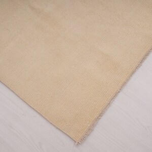 Pu&ograve; includere: Primo piano di un tappeto beige intrecciato con bordo a frangia. Il tappeto &egrave; steso su un pavimento in legno chiaro.