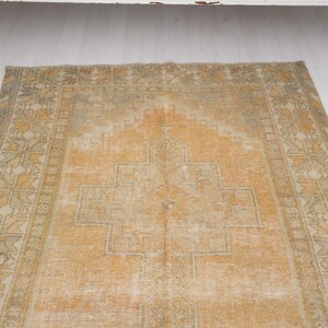 May include: An antique, rectangular rug with a faded, distressed appearance. The rug features a central geometric design in shades of orange and beige, surrounded by a patterned border in similar tones. The rug is placed on a white surface.