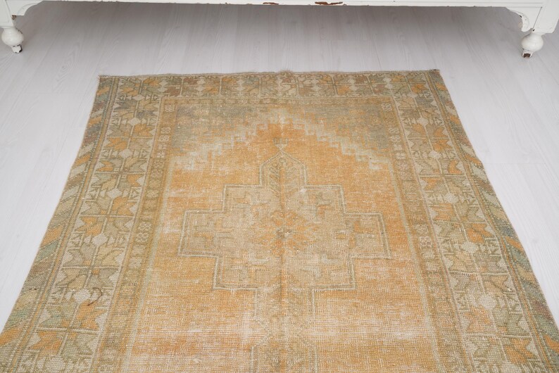 May include: An antique, rectangular rug with a faded, distressed appearance. The rug features a central geometric design in shades of orange and beige, surrounded by a patterned border in similar tones. The rug is placed on a white surface.