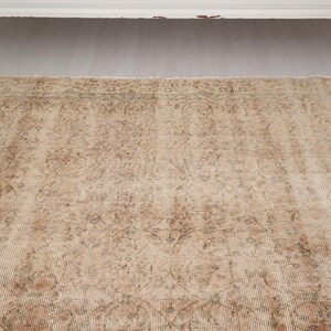 May include: A rectangular, vintage-style rug with a distressed appearance. The rug features a beige and brown color palette with intricate floral and geometric patterns. The rug is placed on a light-colored wooden floor, with a white cabinet in the background.