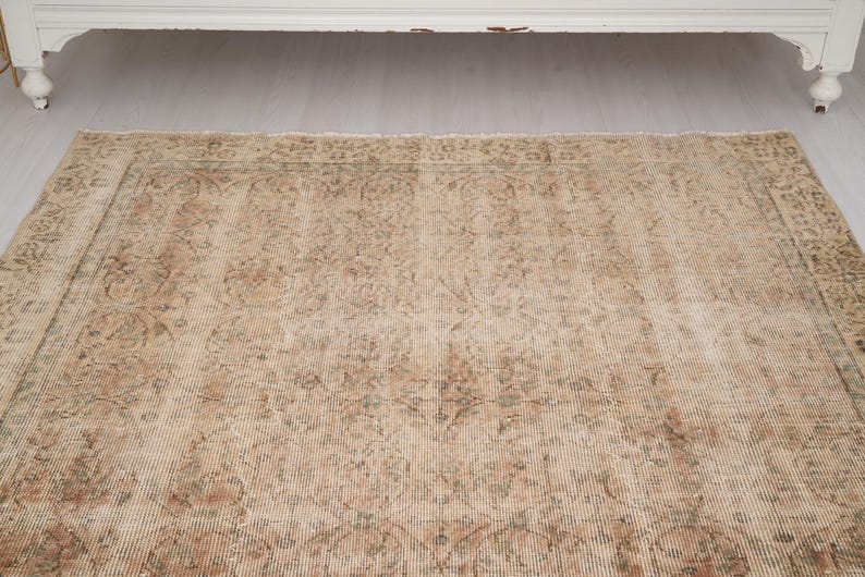 May include: A rectangular, vintage-style rug with a distressed appearance. The rug features a beige and brown color palette with intricate floral and geometric patterns. The rug is placed on a light-colored wooden floor, with a white cabinet in the background.
