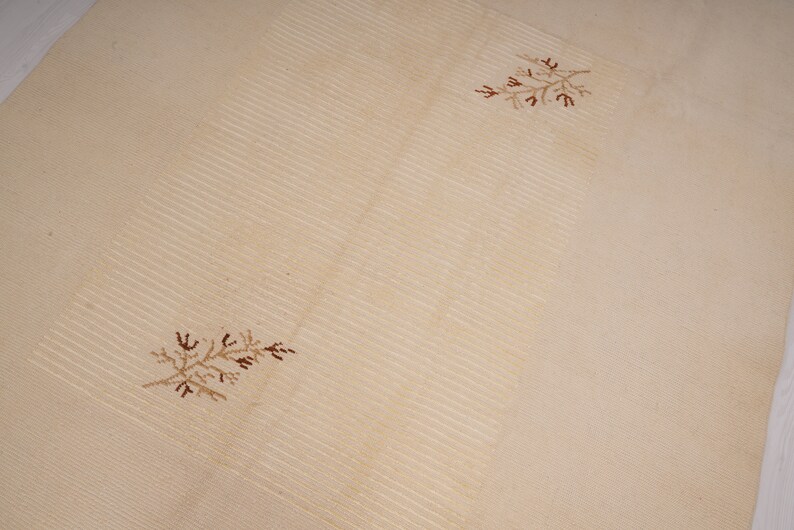 Pu&ograve; includere: Un tappeto beige con una texture leggermente rigata e due disegni floreali ricamati marroni.