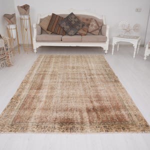 May include: A large, rectangular, vintage-style rug with a faded, distressed appearance. The rug features a central field of beige and brown tones with a detailed border. The rug is placed on a white floor in a room with furniture.