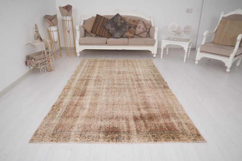 May include: A large, rectangular, vintage-style rug with a faded, distressed appearance. The rug features a central field of beige and brown tones with a detailed border. The rug is placed on a white floor in a room with furniture.