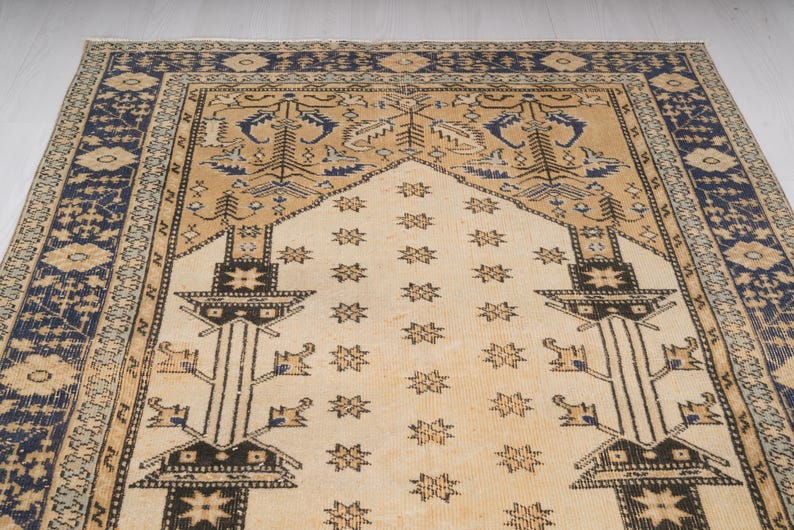 May include: A beige prayer rug with a blue and beige border. The rug has a traditional design with a central motif of a stylized tree and stars.