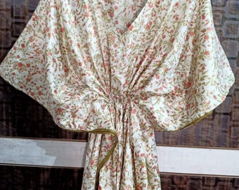 Op de afbeelding: Een witte kaftan met een bloemenprint in tinten groen en oranje. De kaftan heeft een V-hals en korte mouwen. Hij is in de taille geknoopt met een groene sjerp.