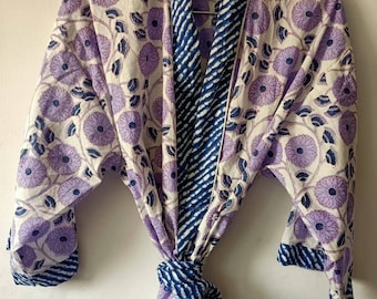 Puede incluir: Una bata de algodón blanca con estampado floral morado y ribete azul a rayas. La bata tiene un cinturón de lazo y mangas largas.