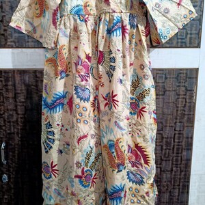 Puede incluir: Un vestido maxi beige con estampado floral con flores azules, rojas y amarillas. El vestido tiene un escote en V y mangas 3/4.