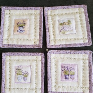 May include: Set of four lavender-themed kitchen pot holders. Each square pot holder features a different lavender design, including a cup of lavender, a field of lavender, and two lavender plants in pots. The pot holders have a white background with a light purple border and a gold embroidered design.