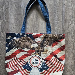 May include: A fabric tote bag featuring a bald eagle graphic centered over a waving American flag background. A circular emblem in the lower center displays the text 250 YEARS. The bag has two blue patterned shoulder straps attached at the top.