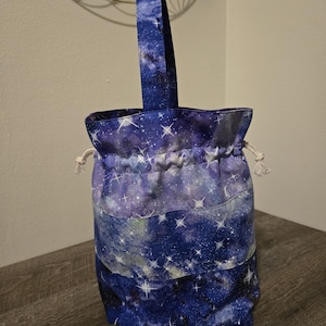 Könnte beinhalten: Ein Kordelzugbeutel mit einem Galaxiedruck in Blau-, Lila- und Weißtönen. Die Tasche hat einen passenden Griff und einen Kordelzugverschluss mit weißem Seil. Die Tasche ist zylindrisch geformt.