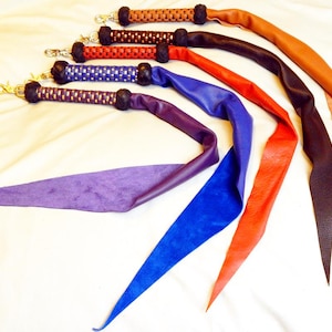 Può includere: Una collezione di cinturini in pelle colorati per borse in varie tonalità, tra cui marrone, rosso, blu e viola. Ogni cinturino presenta un manico intrecciato e una chiusura color oro. I cinturini sono progettati per borse o altri accessori.