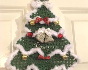 Op de afbeelding: Een handgemaakte gehaakte kerstboomversiering. De groene boom is afgezet met wit, pluizig garen en versierd met rode en gouden bellen, rode strikken en gehaakte bellen. Een grijze ster bekroont de boom. De boom hangt aan een witte deur.