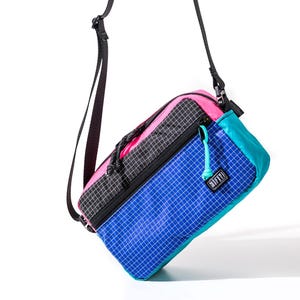 May include: A colorful crossbody bag with a black strap. The bag features a grid pattern in blue, pink, and black. The bag has a zipper closure and a small tag that reads "SIFTYR".
