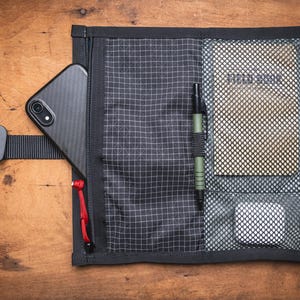 Könnte beinhalten: Schwarze Organizer-Tasche mit Gittermuster, enthält ein Smartphone, einen Stift und einen Notizblock. Die Tasche hat einen schwarzen Riemen mit Schnalle und rote Reißverschlussanhänger. Auf einer Holzoberfläche.