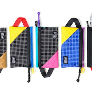 Könnte beinhalten: Fünf farbenfrohe Reißverschlusstaschen mit schwarzen Gittermustern und Akzentfarben. Jede Tasche hat ein anderes Farbschema, darunter Gelb, Blau, Rosa und Grau. Sie haben einen kleinen Griff und einen Karabinerhaken.