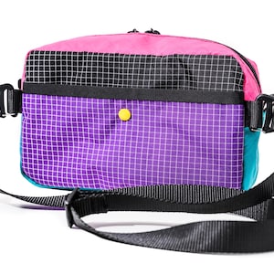 May include: A colorful crossbody bag with a black strap. The bag features a pink top, a black and white checkered section, and a purple grid pattern. A teal side panel and a yellow button add to the design.