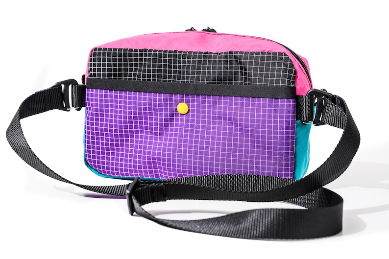 May include: A colorful crossbody bag with a black strap. The bag features a pink top, a black and white checkered section, and a purple grid pattern. A teal side panel and a yellow button add to the design.