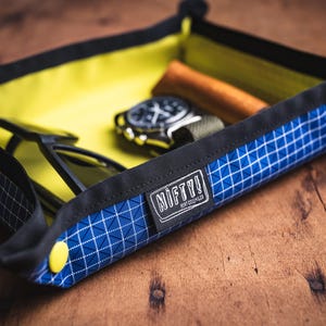 May include: A blue and white patterned tray with a yellow interior holds sunglasses, a watch, and a brown leather item. The tray has a black border and the text "NIFTY!" on a label.