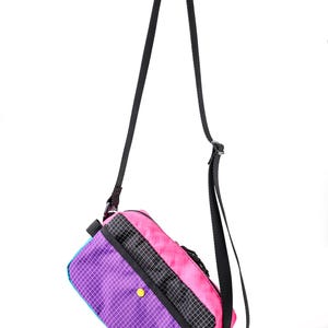 May include: A colorful crossbody bag with a black strap. The bag features a vibrant design with purple, pink, teal, and black panels. A yellow button accents the purple panel. The bag is designed with a grid pattern.