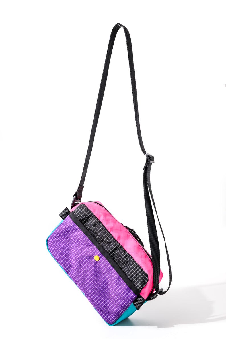 May include: A colorful crossbody bag with a black strap. The bag features a vibrant design with purple, pink, teal, and black panels. A yellow button accents the purple panel. The bag is designed with a grid pattern.
