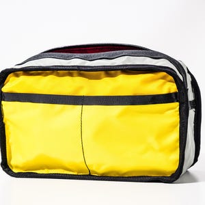 May include: A rectangular travel bag with a bright yellow front and black accents. The bag has a black zipper and a gray top. The interior is a dark red color. The bag is designed for travel.