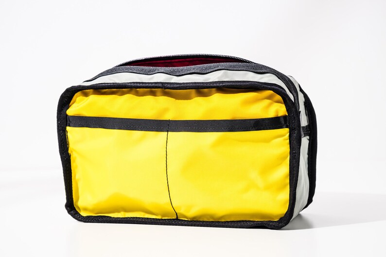 May include: A rectangular travel bag with a bright yellow front and black accents. The bag has a black zipper and a gray top. The interior is a dark red color. The bag is designed for travel.