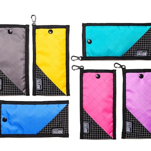 Puede incluir: Una colección de carteras rectangulares de colores en varios tonos, incluyendo gris, amarillo, turquesa, rosa, azul y morado. Cada cartera presenta un patrón de cuadrícula negra y un diseño de bloque de color diagonal. Tienen un cierre a presión y un pequeño mosquetón.