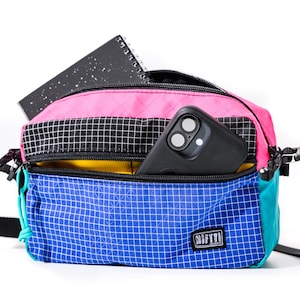 May include: A colorful crossbody bag with a pink top, black and white checkered middle section, and blue bottom. A black smartphone and a black notebook are visible inside. The bag has a black strap and a small logo.