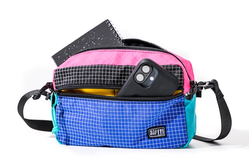 May include: A colorful crossbody bag with a pink top, black and white checkered middle section, and blue bottom. A black smartphone and a black notebook are visible inside. The bag has a black strap and a small logo.