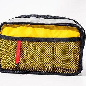 May include: A rectangular travel pouch with a black trim and a yellow interior. The front features a black mesh pocket with three compartments and a red pull tab. The top has a gray panel. The pouch is designed for carrying travel essentials.