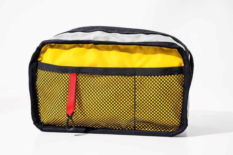 May include: A rectangular travel pouch with a black trim and a yellow interior. The front features a black mesh pocket with three compartments and a red pull tab. The top has a gray panel. The pouch is designed for carrying travel essentials.