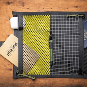 May include: A black zippered pouch with a mesh pocket and a black and white grid pattern. The pouch is open and contains a notebook, a pen, a phone, and a passport.