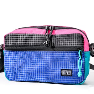 May include: A colorful crossbody bag with a pink top, black center, and blue bottom. The bag has a grid pattern and a teal side. It features a black strap and the word "NIFTY!" on the front.