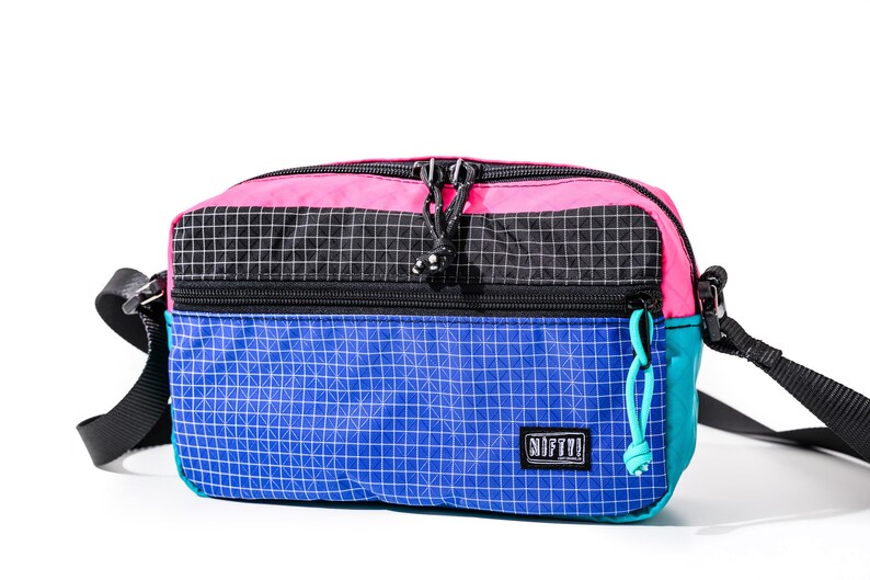 May include: A colorful crossbody bag with a pink top, black center, and blue bottom. The bag has a grid pattern and a teal side. It features a black strap and the word "NIFTY!" on the front.