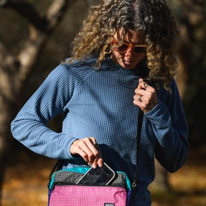 May include: A multi-colored crossbody bag with a pink, teal, black, and purple design. The bag has a checkered pattern on the top and a small rectangular label with the word "RAFFT" on the front. A person is holding a smartphone and putting it into the bag.