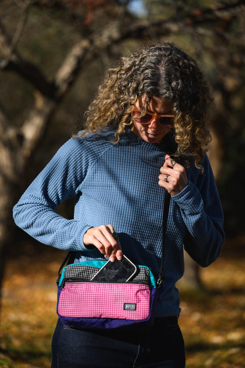 May include: A multi-colored crossbody bag with a pink, teal, black, and purple design. The bag has a checkered pattern on the top and a small rectangular label with the word "RAFFT" on the front. A person is holding a smartphone and putting it into the bag.