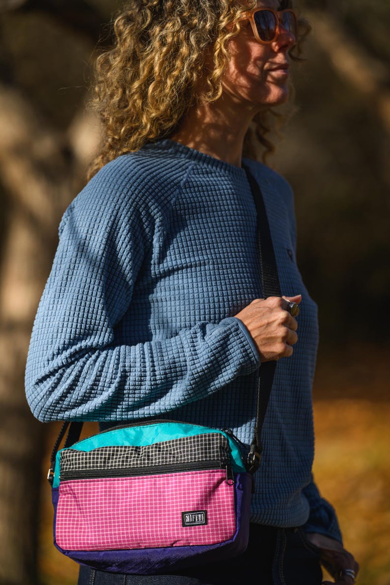 May include: A blue waffle-knit long-sleeve shirt and a multi-colored crossbody bag. The bag features a pink grid pattern, teal, black, and purple panels. The bag has a black shoulder strap and the word "NIFTY" is visible on the bag.