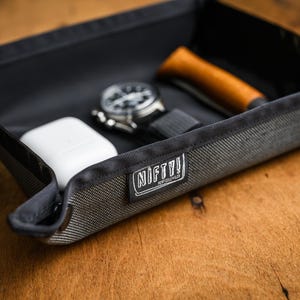 May include: A black, fabric-lined tray with a gray, textured exterior. The tray contains a watch, a small white device, and a wooden tool. The tray has a white label with the text "NIFTY!" printed on it.