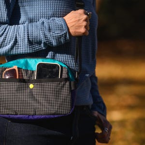 May include: A crossbody bag with a black and white checkered pattern on the front. The bag has a teal top, purple bottom, and a yellow button closure. Sunglasses and a smartphone are visible in the pockets.