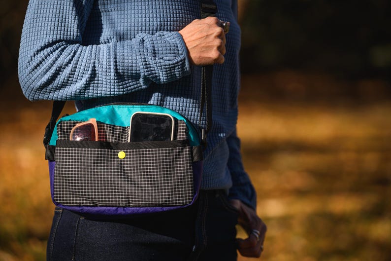 May include: A crossbody bag with a black and white checkered pattern on the front. The bag has a teal top, purple bottom, and a yellow button closure. Sunglasses and a smartphone are visible in the pockets.