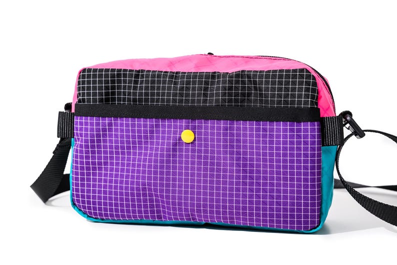 May include: A colorful crossbody bag with a vibrant design. The bag features a pink top, a black and white checkered middle section, and a purple grid-patterned bottom. It has a teal side, a yellow button, and a black strap.