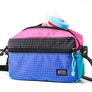 May include: A colorful crossbody bag with a water bottle peeking out. The bag features a pink top, black center, and blue bottom, all with a grid pattern. It has a black strap and a teal zipper pull. The water bottle has a blue cap.
