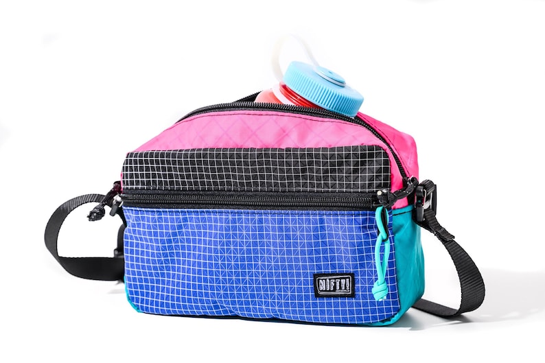 May include: A colorful crossbody bag with a water bottle peeking out. The bag features a pink top, black center, and blue bottom, all with a grid pattern. It has a black strap and a teal zipper pull. The water bottle has a blue cap.