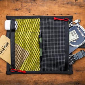 May include: A black zippered pouch with a yellow mesh pocket and a black grid pattern. The pouch is open and contains a notebook, a pen, a passport, a phone charger, and a watch.